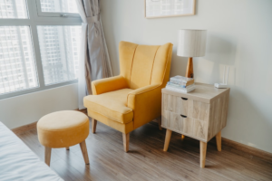 a chair and a footrest next to the dresser with a lamp and books on top